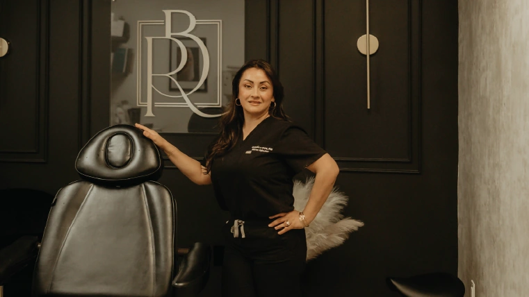 Angelica Alcaraz, BSN, RN, beside the treatment chair at the Revive Beauty Bar logo wall in Salinas